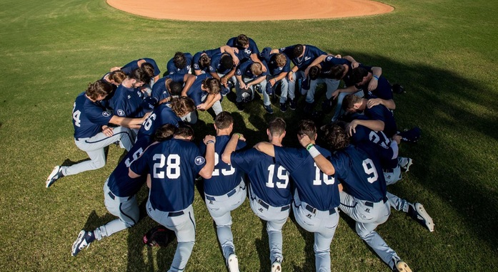 Baseball team huddled together kneeling down with heads bowed on baseball field